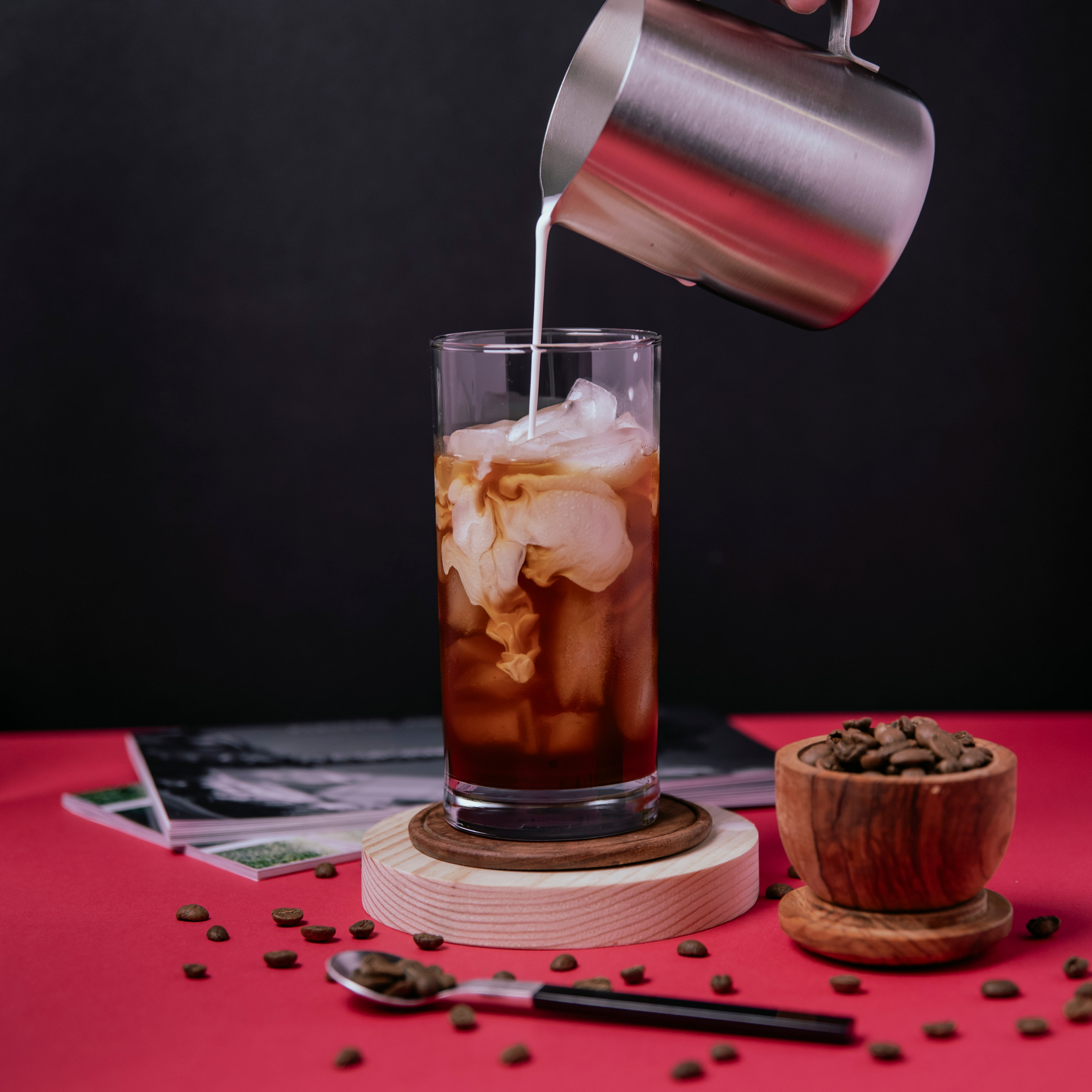 cold brew coffee with bold and sweet flavors pictured on a pink colored surface with a black background near accessories like a coffee spoon and wooden cup holding whole coffee beans. Cream is being poured into the cup of cold brew from a milk frothing container. Casual photo setting